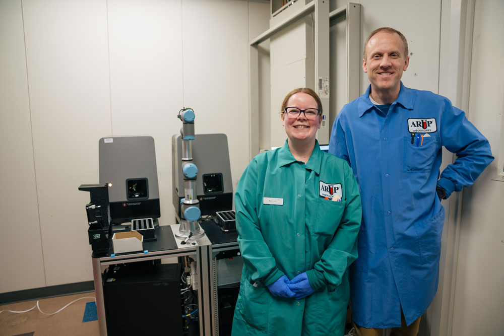 Two people in lab coats stand in front of lab equipment